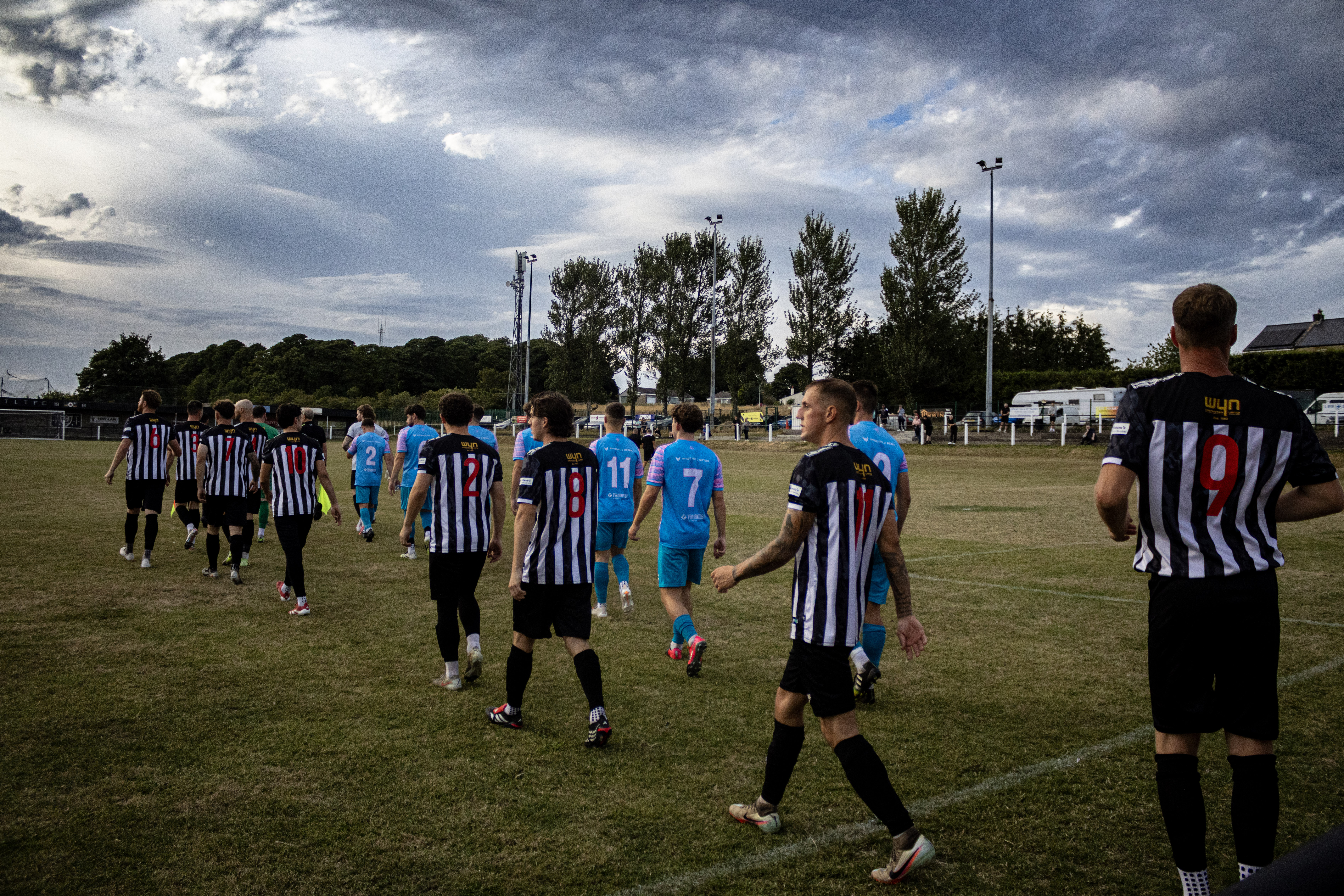 Football teams walking across the pitch beneath a dramatic sky